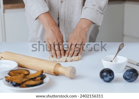 Female hands knead dough on a white table in a bright kitchen. next to it is a rolling pin, a plate with chopped plums, a bowl of cream cheese and a baking dish