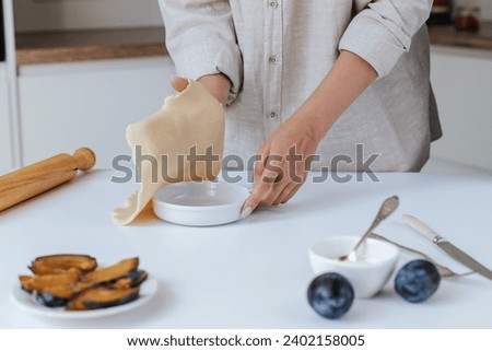 Female hands place dough in a baking dish on a white table in a bright kitchen. next to it is a rolling pin, a plate with chopped plums, a bowl of cream cheese