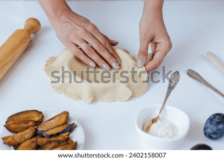 Female hands place dough in a baking dish on a white table in a bright kitchen. next to it is a rolling pin, a plate with chopped plums, a bowl of cream cheese
