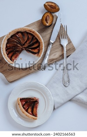 plum pie on a wooden board stands on a white table, next to it are halves of plums and a kitchen towel, a cup of coffee and cutlery. shot from above