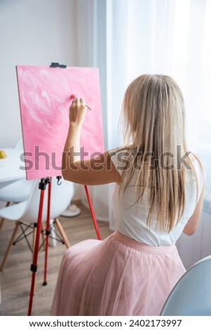 Young blonde woman artist with palette and brush sitting and painting abstract pink picture on canvas near window at home. Art and creativity concept. High quality photo