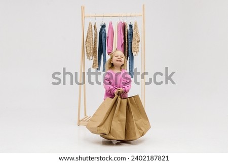 A child, a little girl, stands near the closet, chooses clothes against a light background. Dressing room with clothes on hangers. Wardrobe of children's and stylish clothes. Montessori wardrobe. Royalty-Free Stock Photo #2402187821