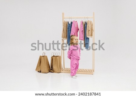 A child, a little girl, stands near the closet, chooses clothes against a light background. Dressing room with clothes on hangers. Wardrobe of children's and stylish clothes. Montessori wardrobe. Royalty-Free Stock Photo #2402187841