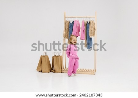 A child, a little girl, stands near the closet, chooses clothes against a light background. Dressing room with clothes on hangers. Wardrobe of children's and stylish clothes. Montessori wardrobe. Royalty-Free Stock Photo #2402187843