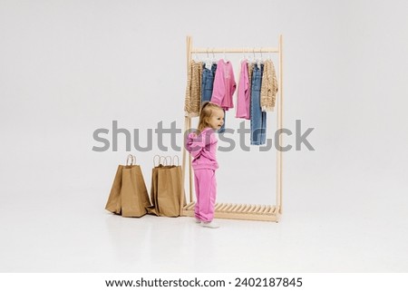 A child, a little girl, stands near the closet, chooses clothes against a light background. Dressing room with clothes on hangers. Wardrobe of children's and stylish clothes. Montessori wardrobe. Royalty-Free Stock Photo #2402187845