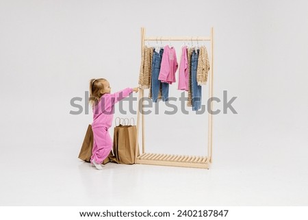 A child, a little girl, stands near the closet, chooses clothes against a light background. Dressing room with clothes on hangers. Wardrobe of children's and stylish clothes. Montessori wardrobe. Royalty-Free Stock Photo #2402187847