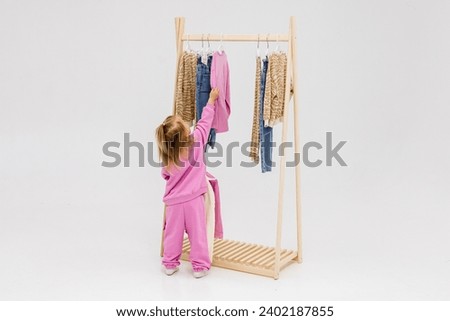 A child, a little girl, stands near the closet, chooses clothes against a light background. Dressing room with clothes on hangers. Wardrobe of children's and stylish clothes. Montessori wardrobe. Royalty-Free Stock Photo #2402187855