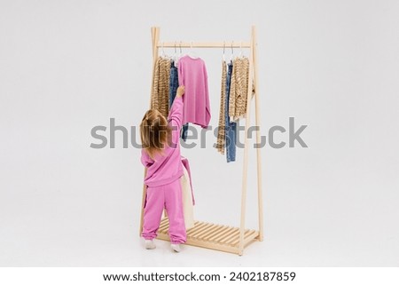 A child, a little girl, stands near the closet, chooses clothes against a light background. Dressing room with clothes on hangers. Wardrobe of children's and stylish clothes. Montessori wardrobe. Royalty-Free Stock Photo #2402187859