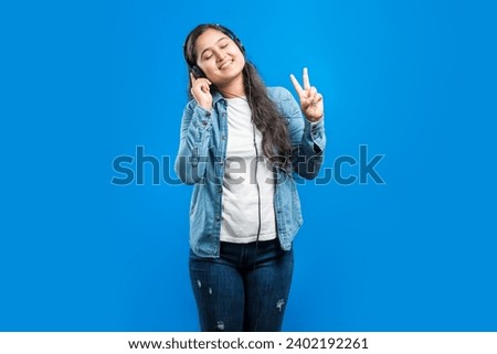 Portrait of a beautiful Indian girl showing v-sign and listening music in her headphones with closed eyes against blue backdrop