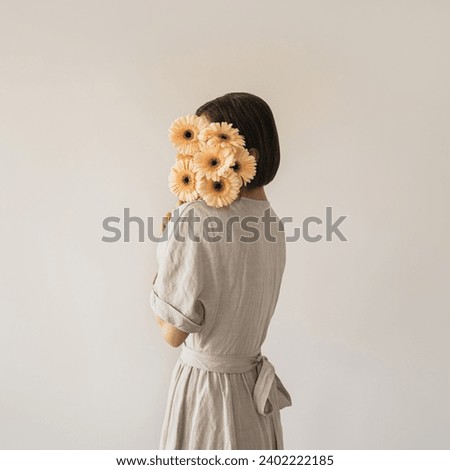 Beautiful young woman in neutral sandy linen dress holding gerber flowers bouquet over white wall. Aesthetic fashion background