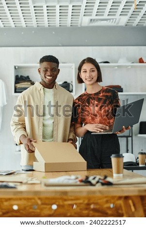 joyful multicultural entrepreneurs with laptop and carton box looking at camera in print studio