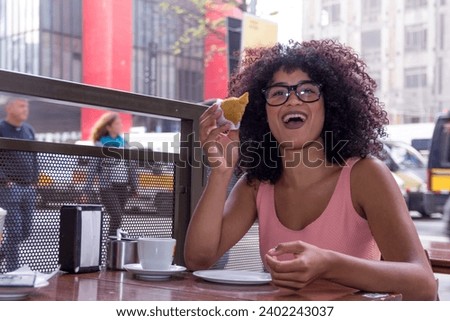 Pretty young woman sitting outside drinking a cup of coffee and eating coxinha food. Typical brazilian snack. In Sao Paulo City during summer period, busy street. Royalty-Free Stock Photo #2402243037