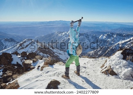 A triumphant tourist at the peak of Mount Toubkal, Morocco, celebrating the ascent with raised hands and an ice axe. Royalty-Free Stock Photo #2402277809