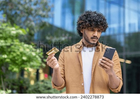 Upset young muslim man standing on city street, holding credit card and looking at phone screen in frustrated, worried way. Royalty-Free Stock Photo #2402278873