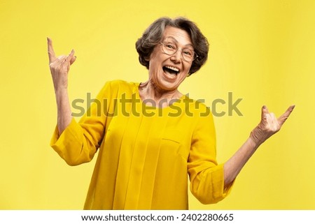 Funny overjoyed senior woman, happy attractive grandmother showing rock sign looking at camera isolated on yellow background. Positive lifestyle, emotions concept 