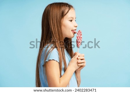 Portrait of positive little girl wearing casual clothes, holding lollipop candy, looking at it, eating, standing isolated on blue background. Concept of desserts, food Royalty-Free Stock Photo #2402283471