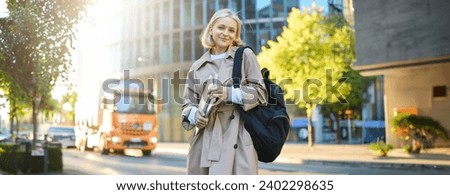 Portrait of smiling, carefree young woman in trench coat, holding backpack and notebook, going to college or university, carries study materials for her language courses.