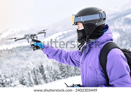 Male snowboarder with equipment and black backpack is launching a flying drone with a remote controller in his hand on winter mountain