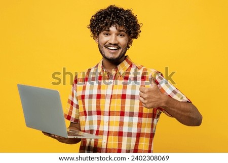 Young smiling happy programmer IT Indian man wears shirt casual clothes hold use work on laptop pc computer show thumb up isolated on plain yellow color background studio portrait. Lifestyle concept