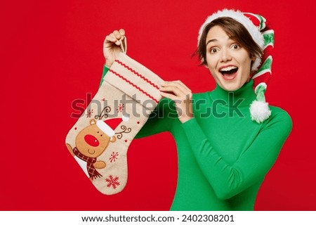 Young surprised amazed excited shocked woman wear green turtleneck Santa hat posing hold stockings with gift isolated on plain red background. Happy New Year 2024 celebration Christmas holiday concept