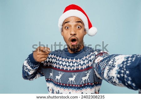 Close up young man wear sweater Santa hat posing doing selfie shot pov on mobile cell phone point finger cmera on you isolated on plain blue background. Happy New Year 2024 Christmas holiday concept