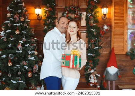 couple with big age difference made a photo shooting for christmas and new year. wooden background with christmas tree. girl in white jeans and peach sweater. man in white shirt and blue jeans