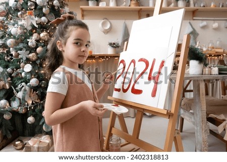 Talented girl artist paints on easel with palette and brush. Christmas tree background. Art school. Little painter.