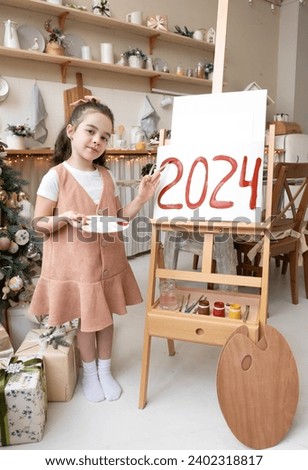Pretty schoolgirl artist paints red numbers 2024 on easel with palette and brush. Christmas tree background. Art school. Happy New Year Eve concept.