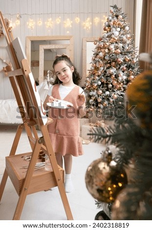 Smiling cute girl paints on easel with palette and paintbrush. Art school. Christmas tree background. Merry Christmas and Happy New Year Eve. Vertical photo.