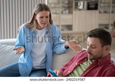 Unpleased wife have quarrel with husband sitting on couch with cellphone at home. Marital discord, emotional couple relationships. Woman try to talk with man ignoring looking at smartphone screen. Royalty-Free Stock Photo #2402319587