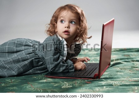 Portrait of an adorable blonde curly hair little girl playing games on a laptop, on gray background, studio shot