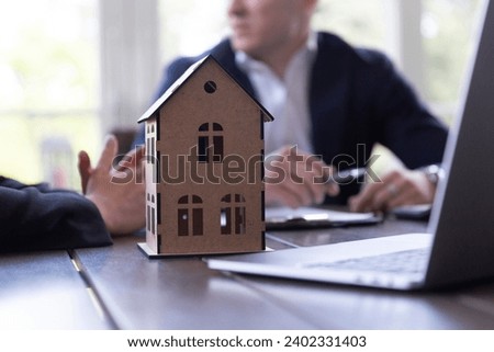 Wooden toy model of house on table in real estate agency office. Businessman and realtor talk about contract signing details