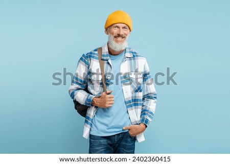 Portrait of handsome mature man wearing stylish casual shirt and yellow hat, holding backpack, standing isolated on blue background. Concept of travel, advertisement