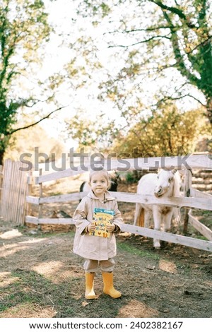 Little girl with a colorful book stands in front of a pony paddock on a farm