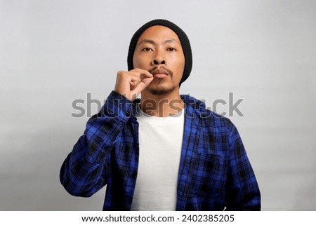 an Asian man, dressed in a casual shirt and wearing a beanie hat, is gesturing for silence by mimicking a zipped mouth, indicating a promise to keep someones secret safe, Isolated on White background