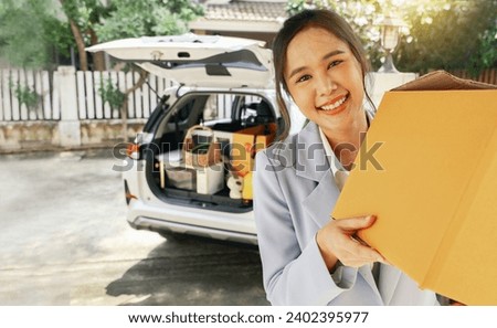 Beautiful asian businesswoman driving car parked in front of her house in a good mood holding cardboard box walking into new residence moving in on a sunny moving day looking happily at the camera. Royalty-Free Stock Photo #2402395977