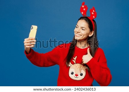 Merry smiling young Latin woman wear red Christmas sweater decorative fun deer horns on head posing do selfie shot on mobile cell phone isolated on plain blue background Happy New Year holiday concept
