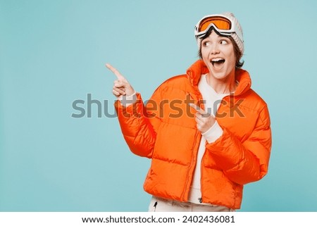 Skier young woman wearing warm padded windbreaker jacket hat ski goggles mask point index finger aside on area travel rest spend weekend winter season in mountains isolated on plain blue background