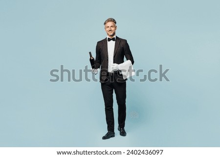 Full body adult sommelier barista male waiter butler man wears shirt black suit bow tie uniform hold in hand bottle of wine work at cafe isolated on plain blue background. Restaurant employee concept Royalty-Free Stock Photo #2402436097