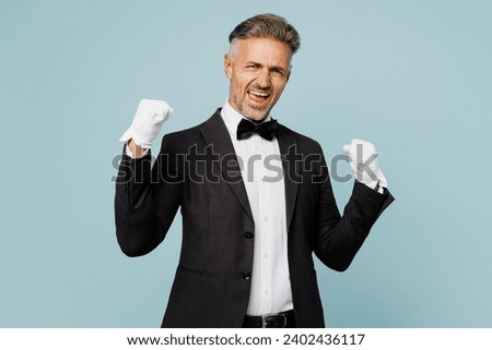 Adult barista male waiter butler man wear shirt black suit bow tie elegant uniform do winner gesture celebrate clench fists work at cafe isolated on plain blue background. Restaurant employee concept