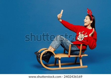 Full body merry young Latin woman wear red Christmas sweater decorative fun deer horns on head sledding doing selfie shot on mobile cell phone isolated on plain blue background. Happy New Year concept
