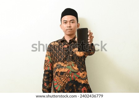 portrait of an Asian man in batik shirt and cap holding and displaying a smartphone screen. Indonesian Muslim man showing cell phone screen on isolated white background