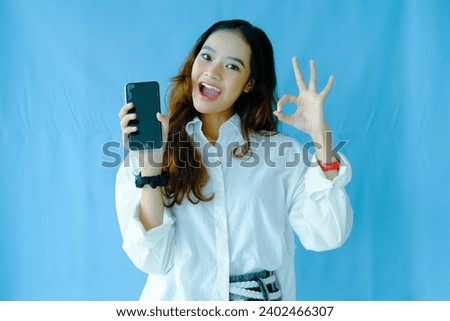 Happy young Asian woman showing OK sign and using mobile phone on blue background