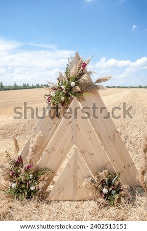 Wedding wooden arch decorated with a detailed flower arrangement in a golden, mown field under blue summer sky. Rustic style.