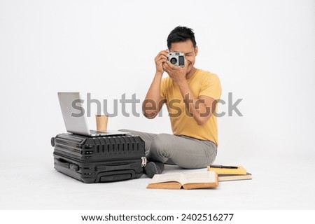 A positive Asian man is preparing his luggage for his holiday trip, checking his camera, sitting on an isolated white studio background.