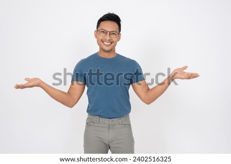 A positive Asian man in casual clothes is opening his palm widely, smiling at the camera, standing on an isolated white background. showing option, recommendation, presentation, friendly welcome Royalty-Free Stock Photo #2402516325
