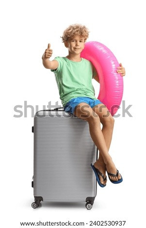 Boy with a swimming ring sitting on a suitcase and gesturing thumbs up isolated on white abackground