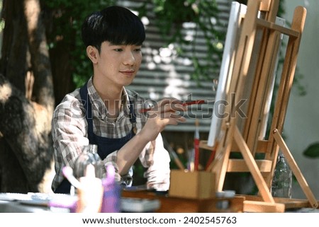 Handsome Asian male wearing apron painting picture with brush on easel in the garden.