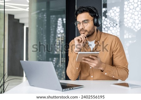 Thinking concentrated man studying sitting inside office at workplace, businessman watching video training course, jotting down data in notebook, using laptop, employee upgrading skills, video call. Royalty-Free Stock Photo #2402556195