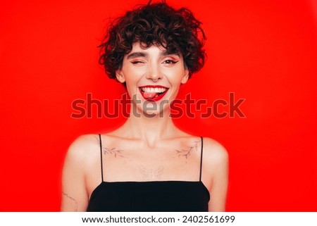 Young beautiful smiling female in trendy summer black tank top. Carefree woman posing near red wall in studio with curly hairstyle. Positive model having fun. Cheerful and happy. Shows tongue